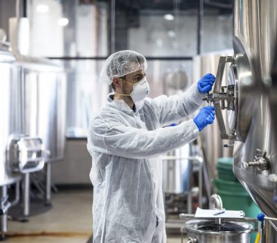 Technologist in protective uniform wearing hairnet mask goggles and gloves closing industrial tank reservoir.